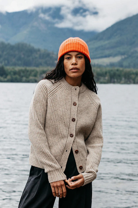 Woman wearing an orange beanie and beige cardigan standing by a lake with mountains in the background