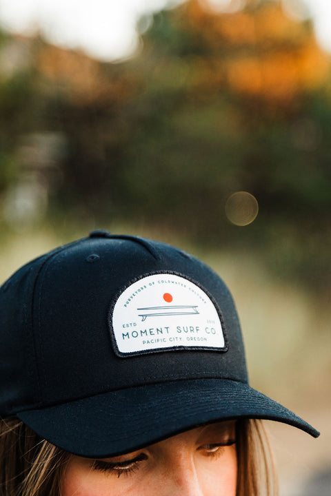 Person wearing a navy blue cap with a white patch featuring text and a red dot.