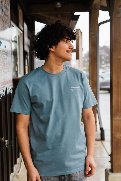 Person wearing a light blue t-shirt with a logo, standing outdoors.