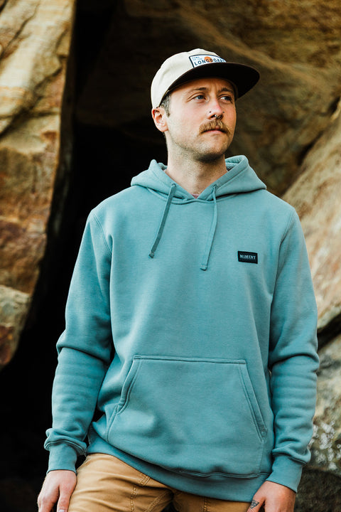 Man wearing a teal hoodie and cap standing against a rocky background