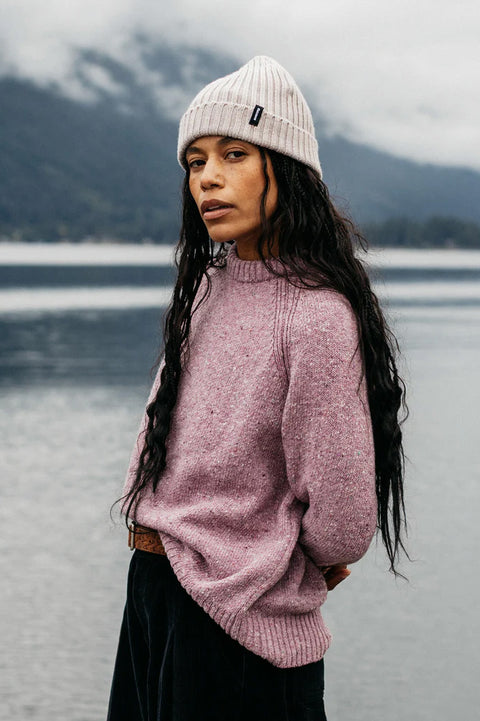 Person wearing a pink sweater and white beanie standing by a lake with mountains in the background
