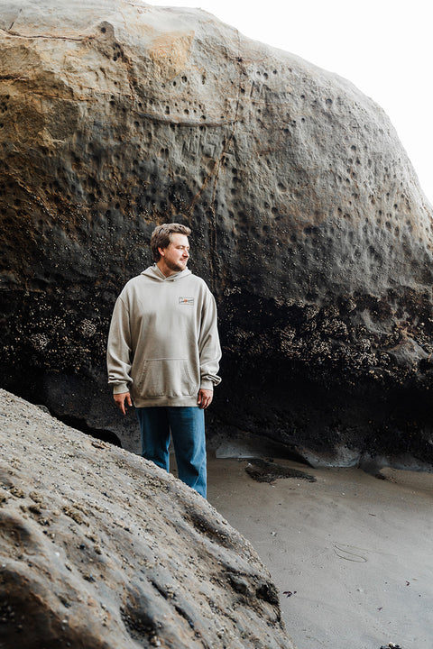 Man standing on a beach with large rocks in the background wearing the Moment The Classic Pullover Hoodie - Faded Taupe