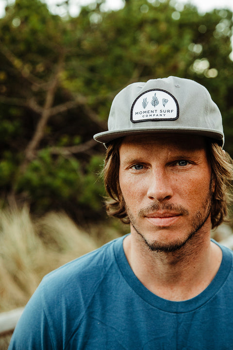 A closeup of a male model looking right at the camera wearing the Moment Kelp Friends 5 Panel Hat.