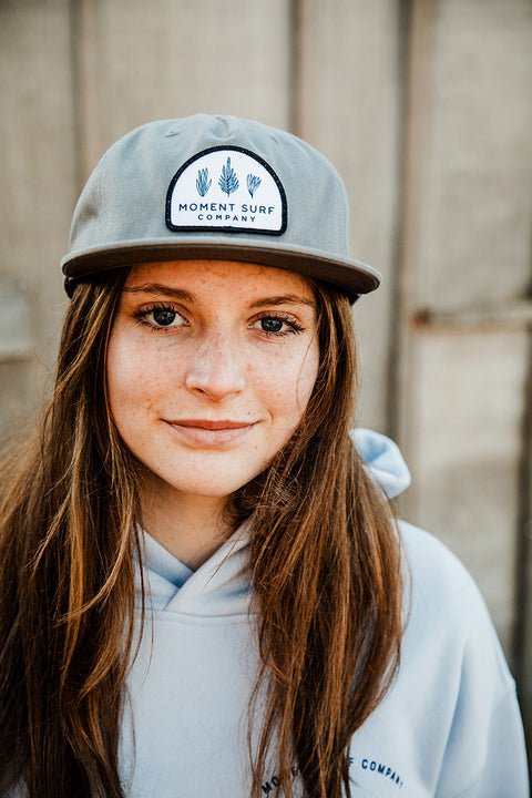 A female model looking right at the camera wearing the Moment Kelp Friends 5 Panel Hat.