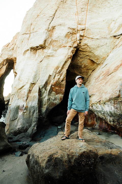 Person standing on a rock with large rock formations in the background wearing the Moment Ridge Pullover Hoodie - Lake Green