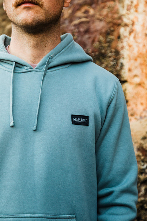 Person wearing a teal hoodie with a Moment logo against a textured wall.