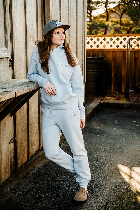 A model wearing the Moment Women's Lt. Dan Sweatpant leaning up against a wood shelf.