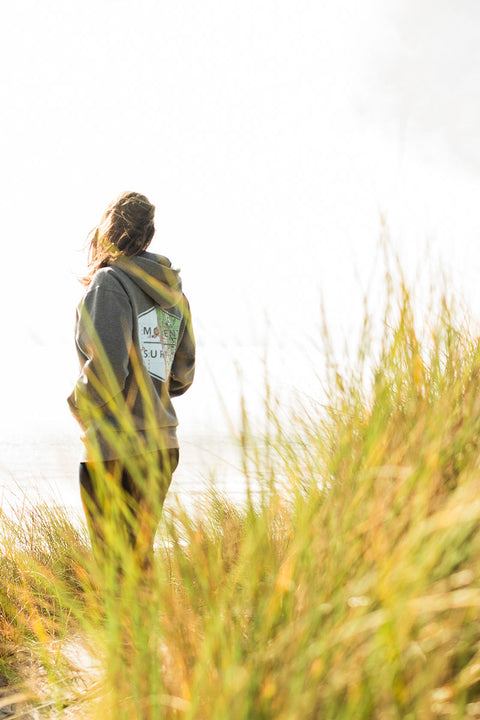 A woman wearing the Moment Boxed Topo Logo Pullover Hoodie - Gunmetal Heather at the beach looking at the ocean.